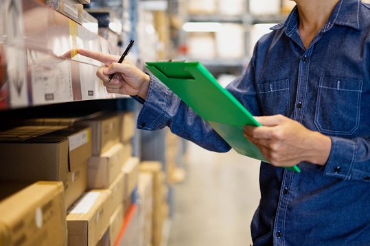 Someone in a warehouse holding a clipboard and pointing at a box with the implications of taking inventory