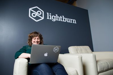 Nora Lahl seated under a lit Lightburn sign, working on her laptop and smiling 