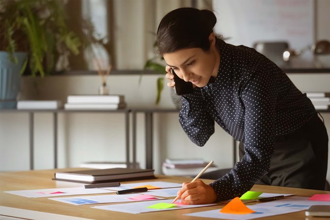 A designer on a phone call standing over papers and sticky notes and taking notes with a pencil