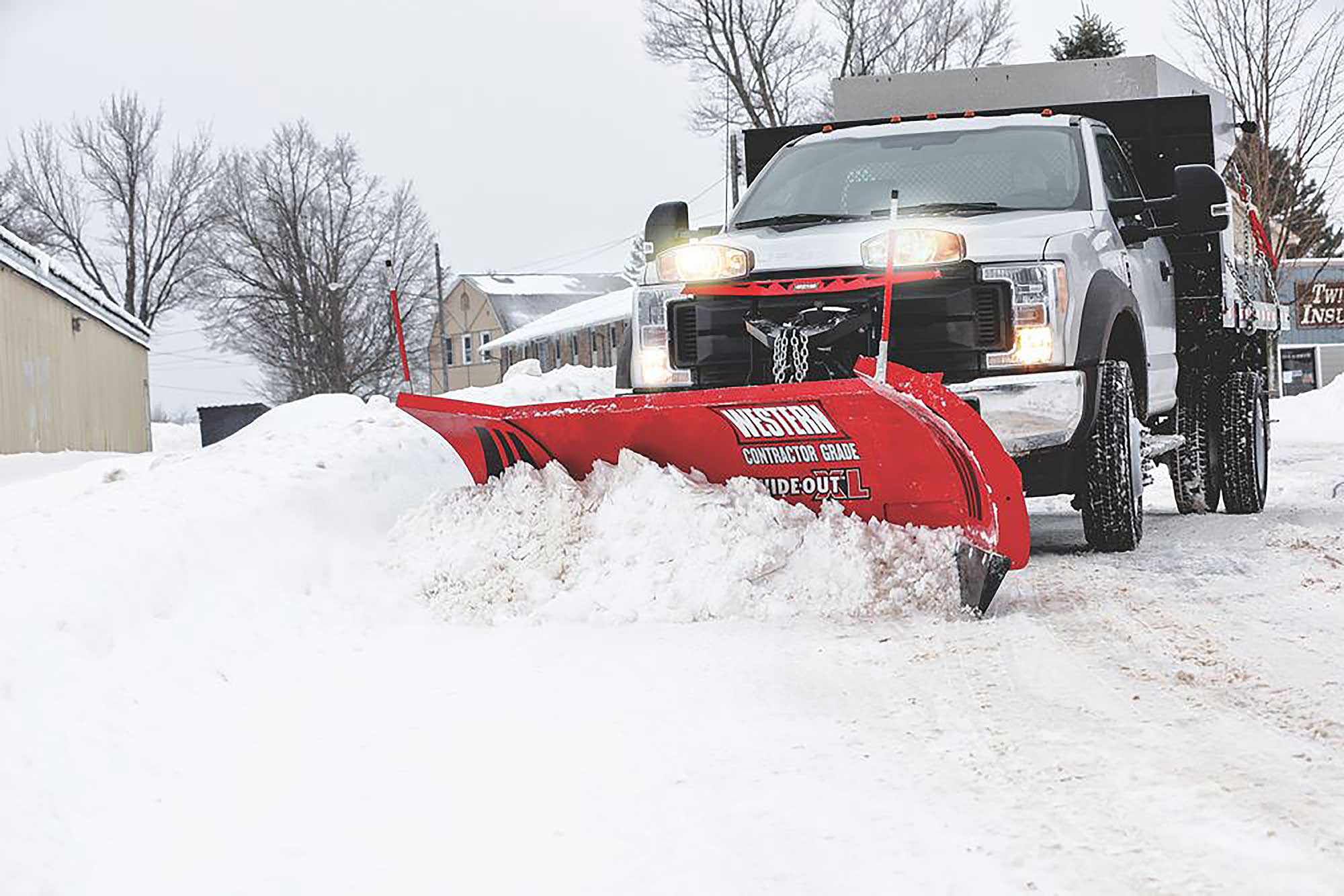 White heavy-duty dump truck with headlights on, fitted with a red Western Contractor Grade Wide-Out XL snow plow, clearing snow from a road on an overcast winter day with bare trees and buildings in the background.