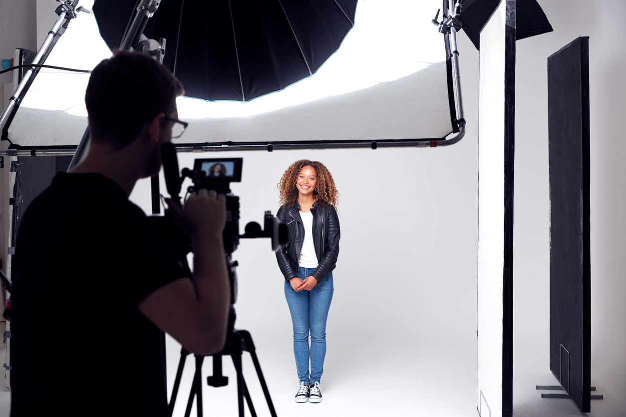 A photographer behind a tripod-mounted camera shoots a smiling young woman in a leather jacket and jeans posing against a white backdrop in a professional studio.