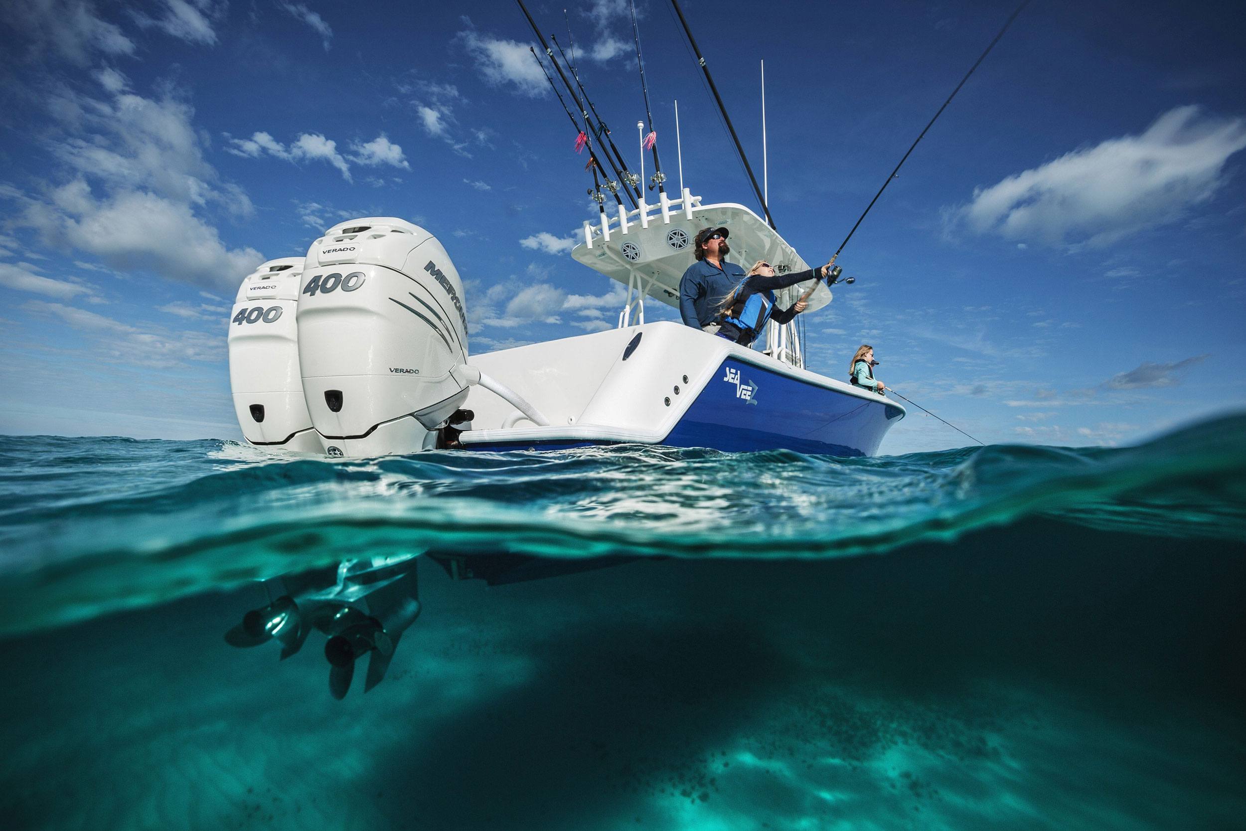 A family fishes from a SeaVee center-console boat powered by twin Mercury Verado 400 outboard motors, shot from a split-level water angle showing the props beneath the clear turquoise surface.